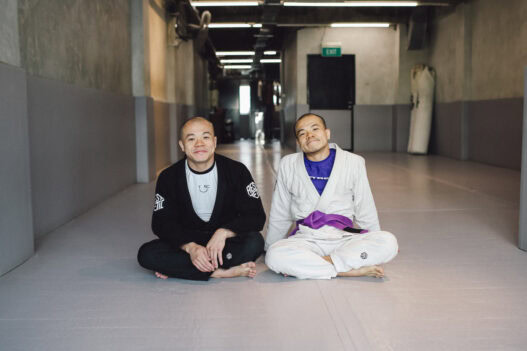 Two men in Brazilian Jiu-Jitsu gis sitting on the mat in a martial arts training facility.