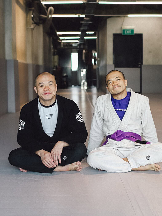 Two men in Brazilian Jiu-Jitsu gis sitting on the mat in a martial arts training facility.