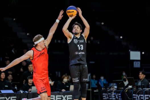 An image of a basketball player in black uniform shooting the ball while an opponent in red attempts.