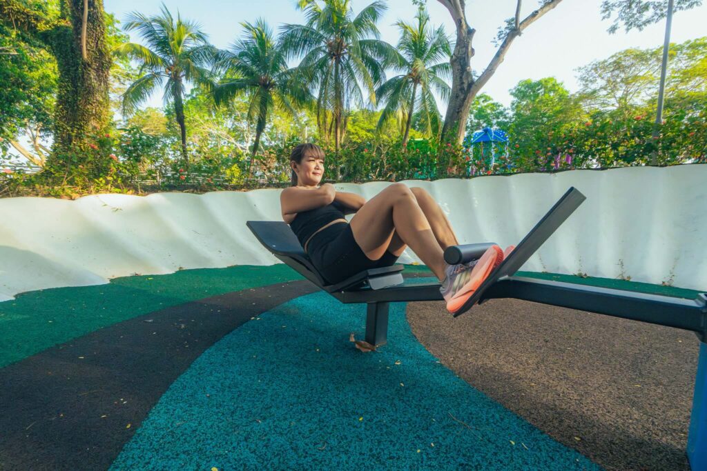 Sit-up exercise on outdoor fitness equipment in a lush, tropical park setting with palm trees and vi.